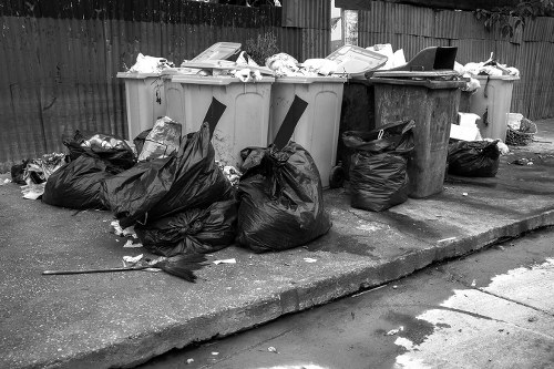 A row of large wheelie bins and several black rubbish bags are positioned on a paved driveway, likely part of a house clearance process in Bracknell Forest. The bins are made of plastic, with some in light grey and one in dark grey, featuring hinged lids that are partially open or closed, with various paper and cardboard waste visible on top of or near them. The black rubbish bags are crumpled and stacked close to the bins, some leaning against the bins and others on the ground. The surface underneath is concrete, showing typical wear, with minor cracks visible. In the background, a wooden fence provides enclosure, and the environment appears to be an outdoor residential area. The scene is lit naturally, with diffuse lighting suggesting an overcast day, congruent with waste collection activities managed by services like House Clearance Bracknell Forest for efficient rubbish disposal and clearance in the Bracknell area, with a typical urban setting in Berkshire.