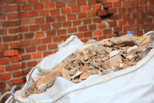 A close-up view of a large white sack filled with mixed rubble and building debris, including broken red bricks, fragments of concrete, and small wooden sticks, situated outdoors on a paved surface in front of a brick wall. The debris appears freshly gathered, with sharp-edged pieces and varying textures. The scene likely depicts a waste collection process being carried out by House Clearance Bracknell Forest, a local rubbish removal service operating in the Bracknell Forest area. The background features a red brick wall, providing a typical outdoor setting, and the bag's material is crinkled and slightly stained from handling. The photograph emphasizes the physical nature of rubbish removal, showcasing the types of waste materials that are commonly cleared during house or site clearance projects in the region.