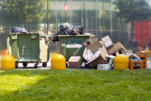 A row of three large waste bins and numerous cardboard boxes and bags overflowing with discarded items are positioned on a paved area in front of a reflective glass building, with a well-maintained grassy verge in the foreground. The bins are green and made of plastic, with the lids open or partially covered by black rubbish bags. Cardboard boxes of various sizes, some flattened and others still assembled, are scattered on the ground alongside plastic crates and bags which contain miscellaneous waste. The scene appears to be outside a commercial or residential property, possibly in the Bracknell Forest area, with trees and a red sign visible in the background, and the sunlight casting shadows indicating daytime. The presence of these waste materials aligns with rubbish removal services such as those offered by House Clearance Bracknell Forest, emphasizing the need for professional clearing and disposal in the local area.