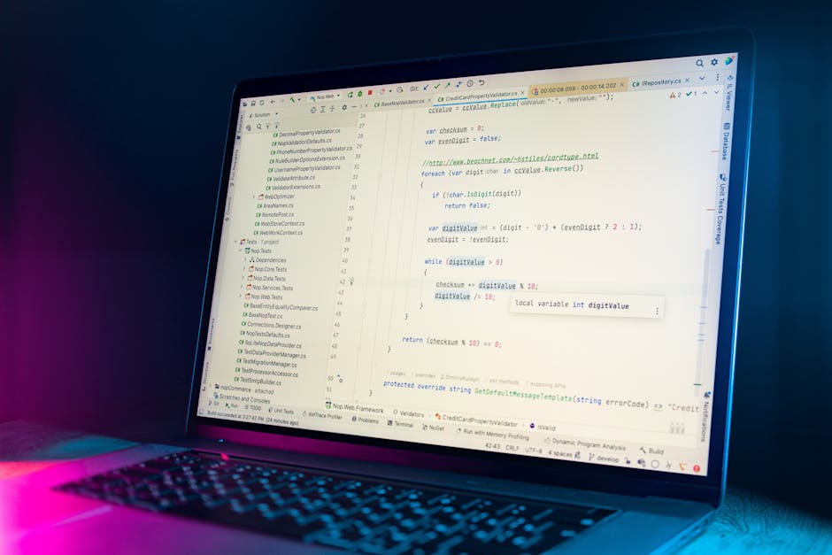 A close-up image of a person's right hand, wearing a black wristwatch, typing on a laptop keyboard placed on a wooden surface. The laptop's screen displays lines of programming code with highlighted s