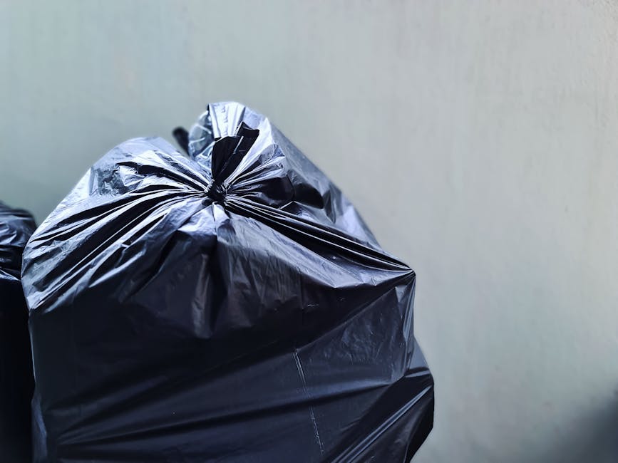 Close-up image of a tightly tied black plastic rubbish bag, positioned towards the foreground on the left side of the frame. The bag has a glossy surface with visible creases and folds, indicating it 